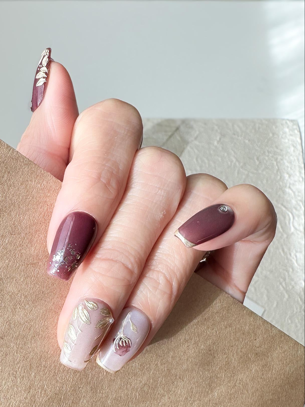 Hand with press-on nails in square coffin shape in deep plum and mauve tones with gold leaf accents, metallic floral designs, and silver gemstone details.