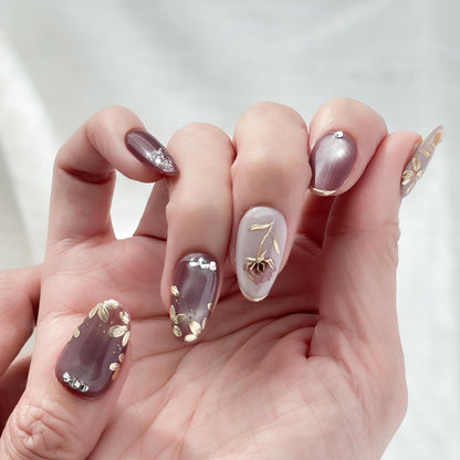 Hand with almond shaped Press-on nails in deep plum and mauve tones with gold leaf accents, metallic floral designs, and silver gemstone details