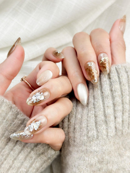 Close-up of almond-shaped nude and caramel brown press-on nails with gold foil accents and subtle shimmer, styled against a soft beige knit sweater.