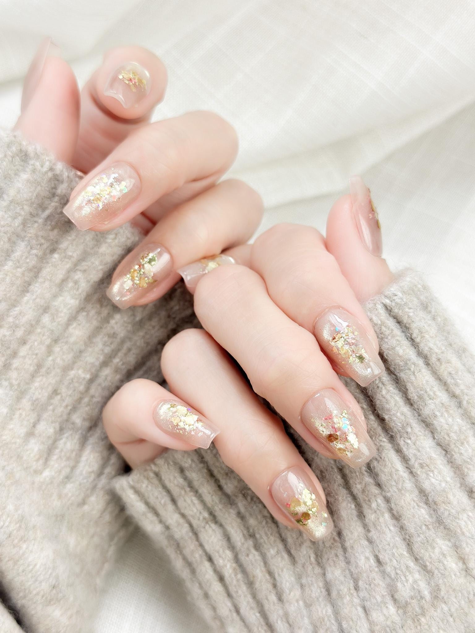 Close-up of hands with nude nail polish and gold glitter on a textured beige background, press on nails
