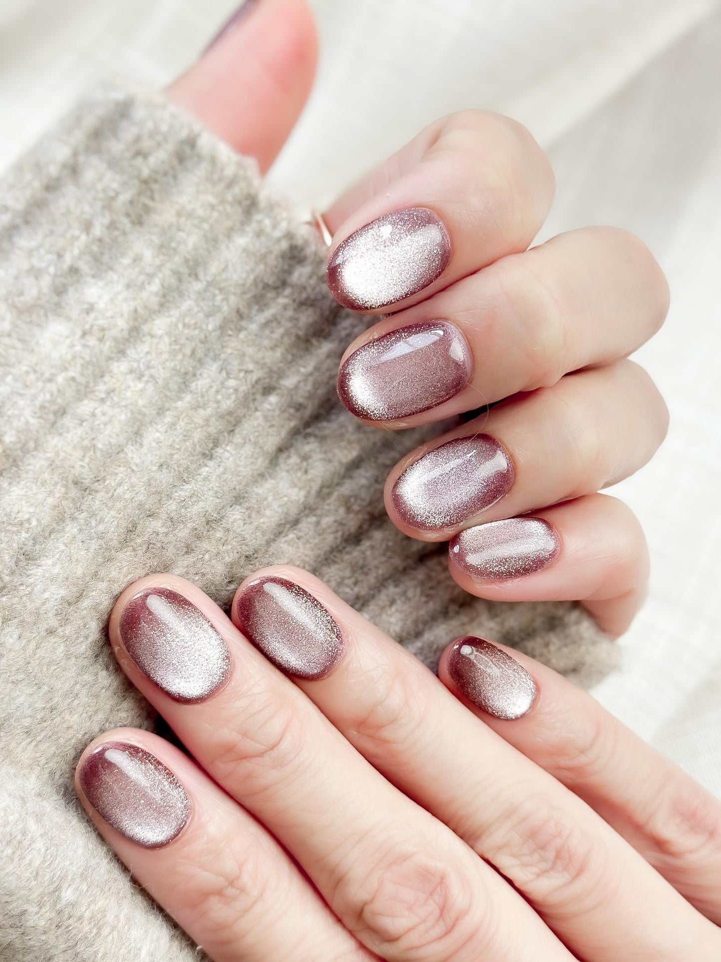 Short oval press-on nails in a metallic mauve cat-eye shimmer, glossy finish, luxury reusable press-on manicure by ManiFleur. Elegant mauve shimmer nails styled against a cozy knit sweater