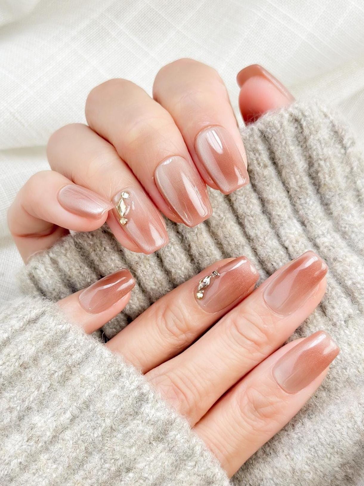 Close-up of a hand wearing glossy nude-brown press-on nails with a subtle cat-eye shimmer, featuring a gold leaf and crystal accent on one nail. The manicure is styled against a soft gray knit sweater for an elegant, cozy look