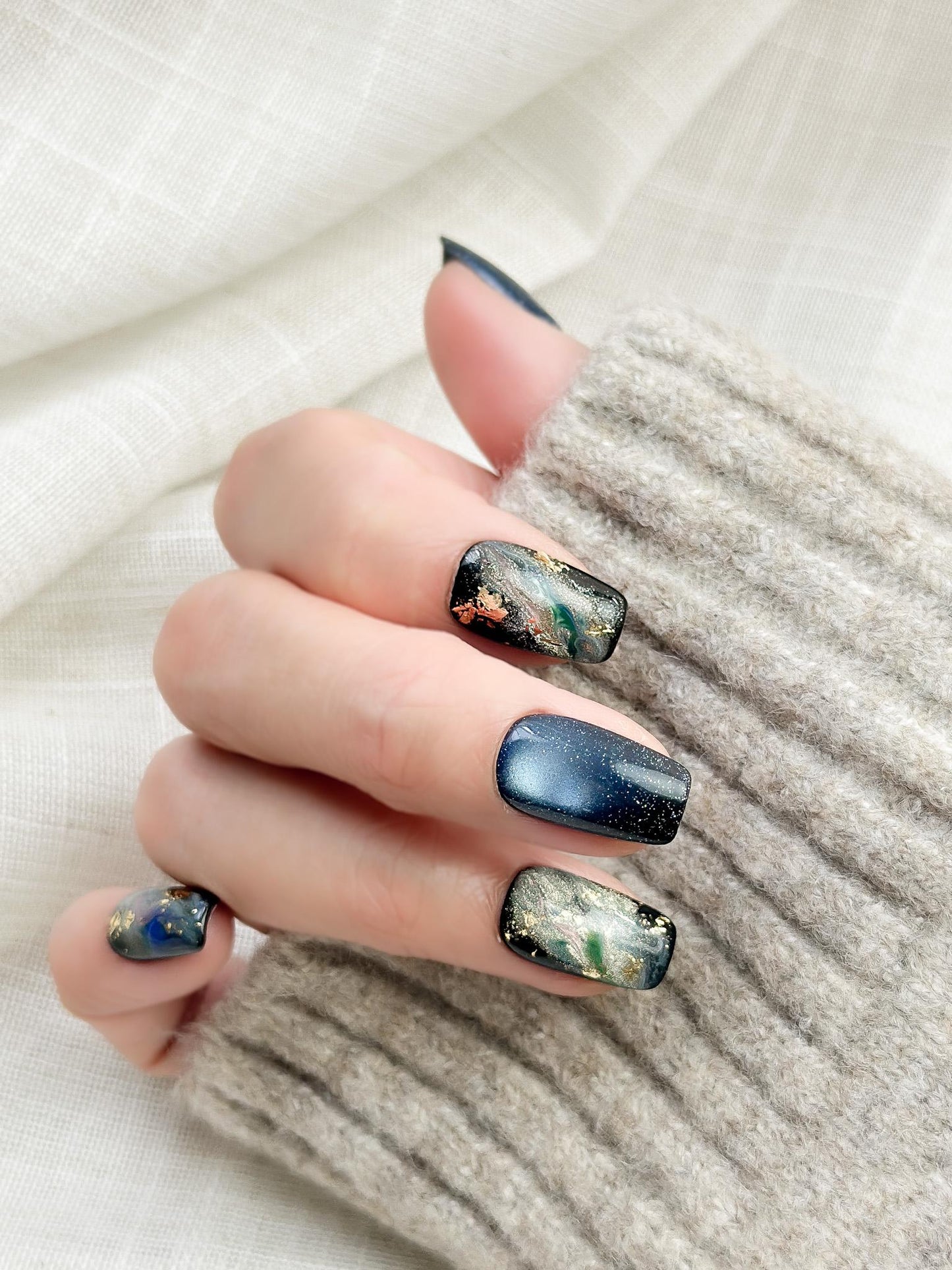 Hand with blue cat-eye press-on nails with galaxy shimmer and abstract marble accents in green, gold, and silver foil