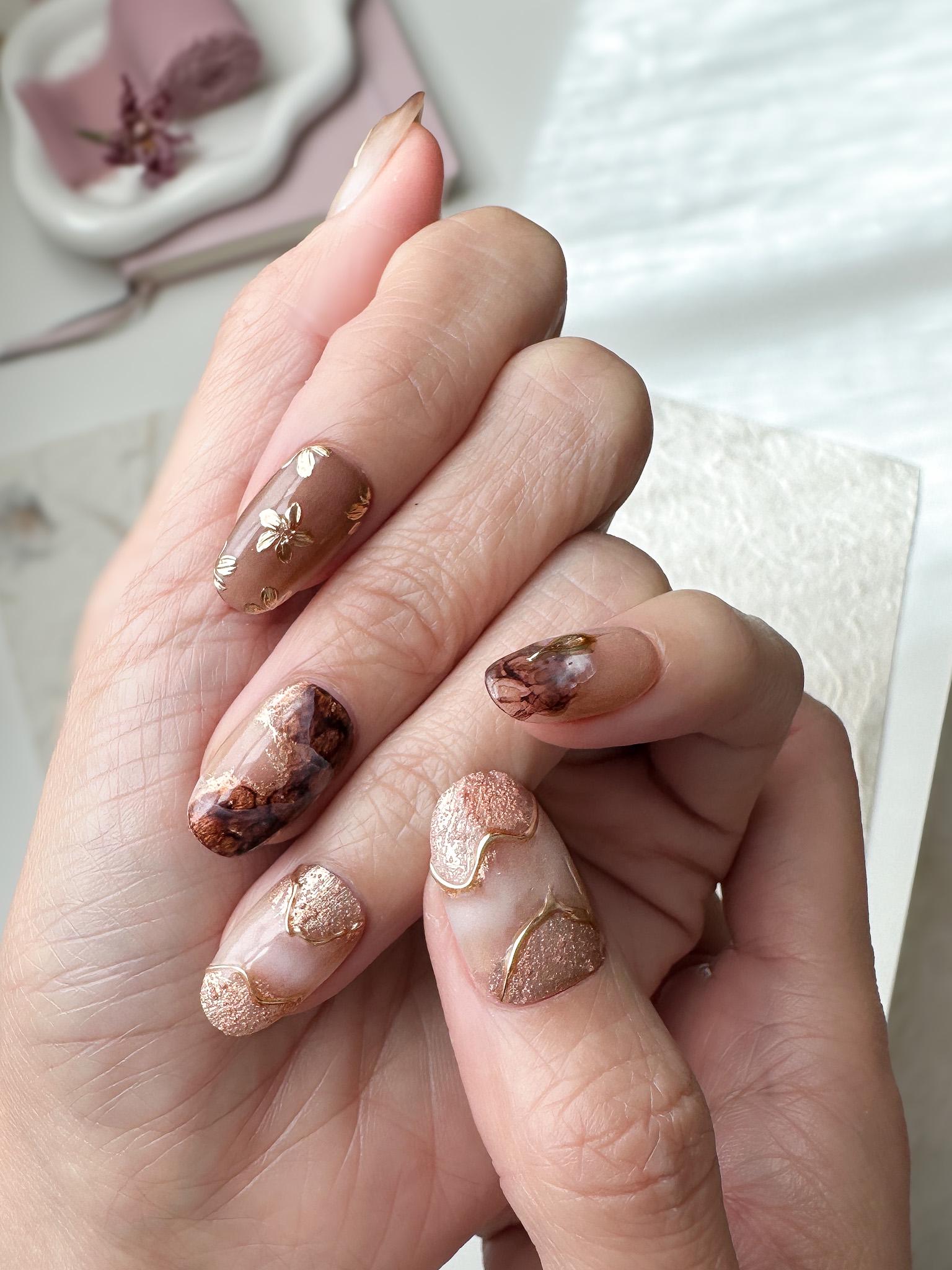 Hand with press-on nails in neutral tortoise design and mocha tones with gold foil accents, marble detailing, and embossed metallic floral designs