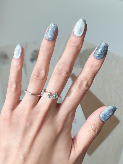 Hand with Blue and silver marble press-on nails with icy shimmer, metallic accents, and scattered silver foil flakes. Winter-inspired design featuring frosted blue hues and glossy snow-like details