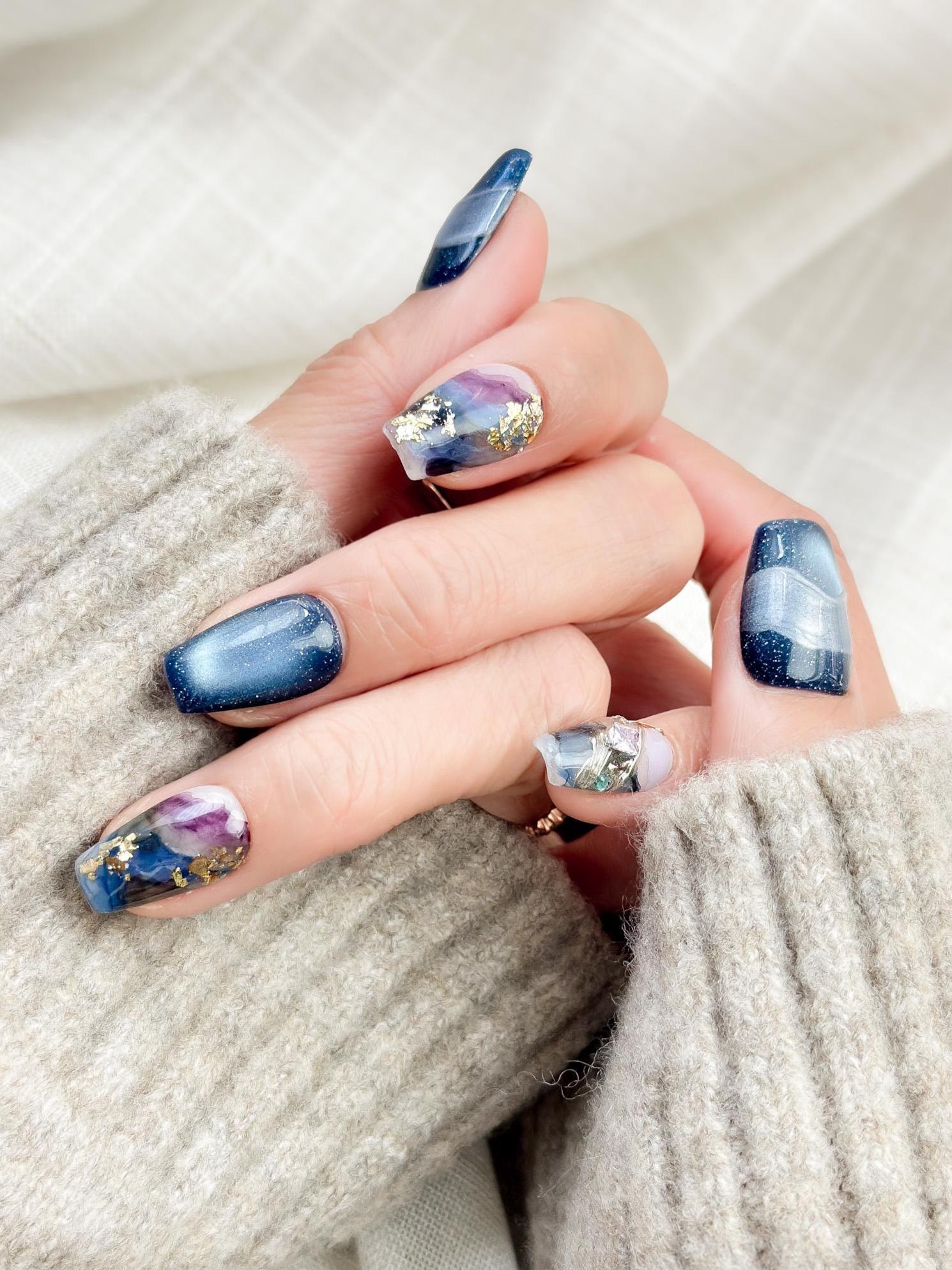 Hand with Blue galaxy press-on nails with starry cat-eye shimmer and watercolor marble accents in navy, lavender, and white, finished with gold foil details