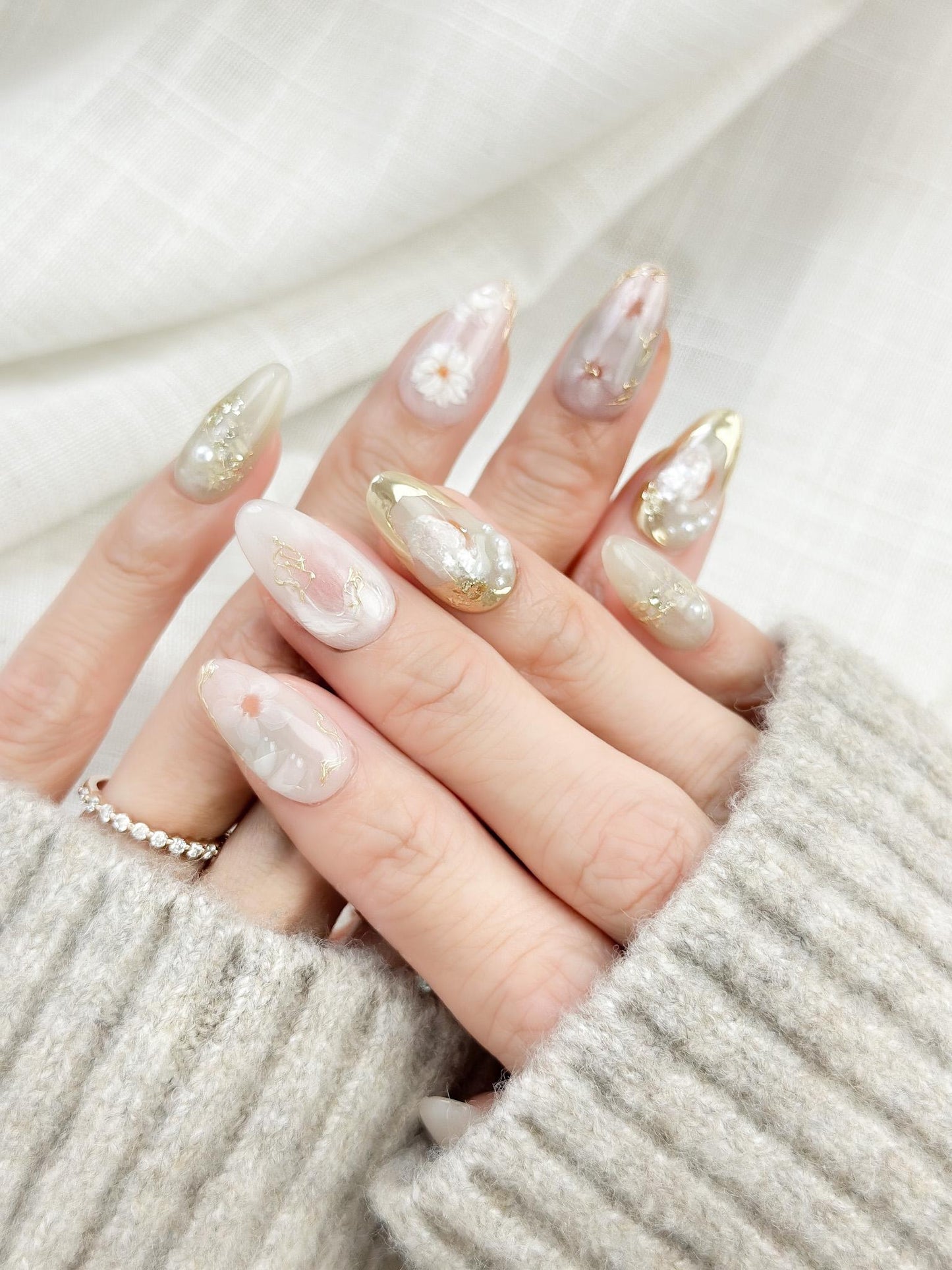 Hand with Almond-shaped white and pearlescent press-on nails with 3D floral accents, gold chrome details, pearl embellishments, and soft milky marble art