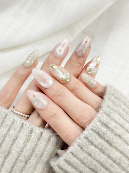 Hand with Almond-shaped white and pearlescent press-on nails with 3D floral accents, gold chrome details, pearl embellishments, and soft milky marble art