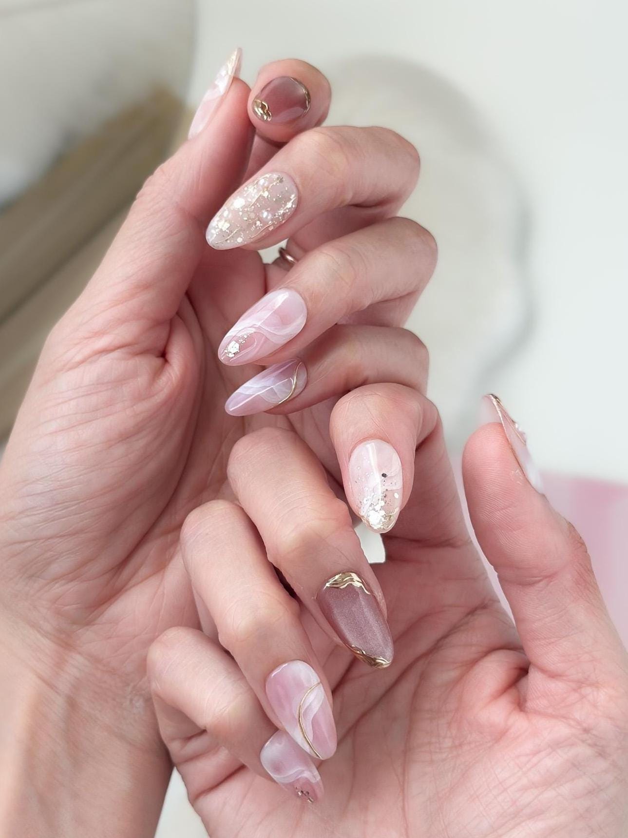 Close of of hand with Pink and nude luxury press-on nails with marble swirl designs, gold metallic accents, pearls, and glitter