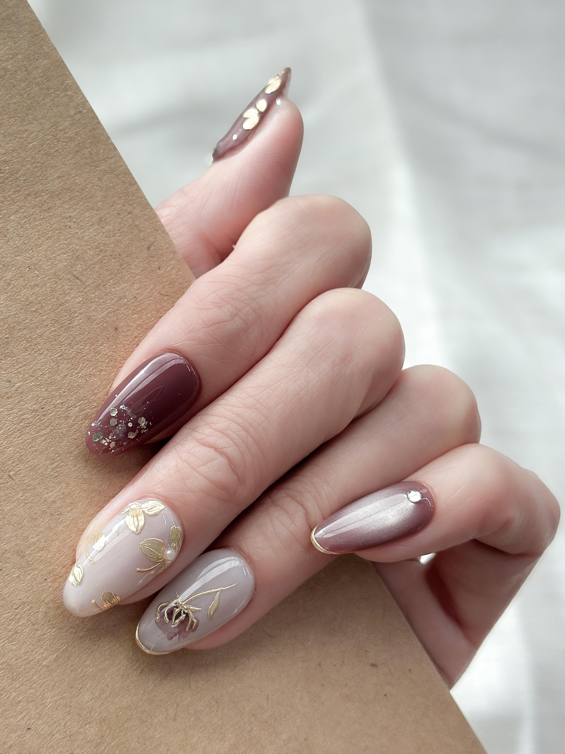 Hand with almond shaped Press-on nails in deep plum and mauve tones with gold leaf accents, metallic floral designs, and silver gemstone details