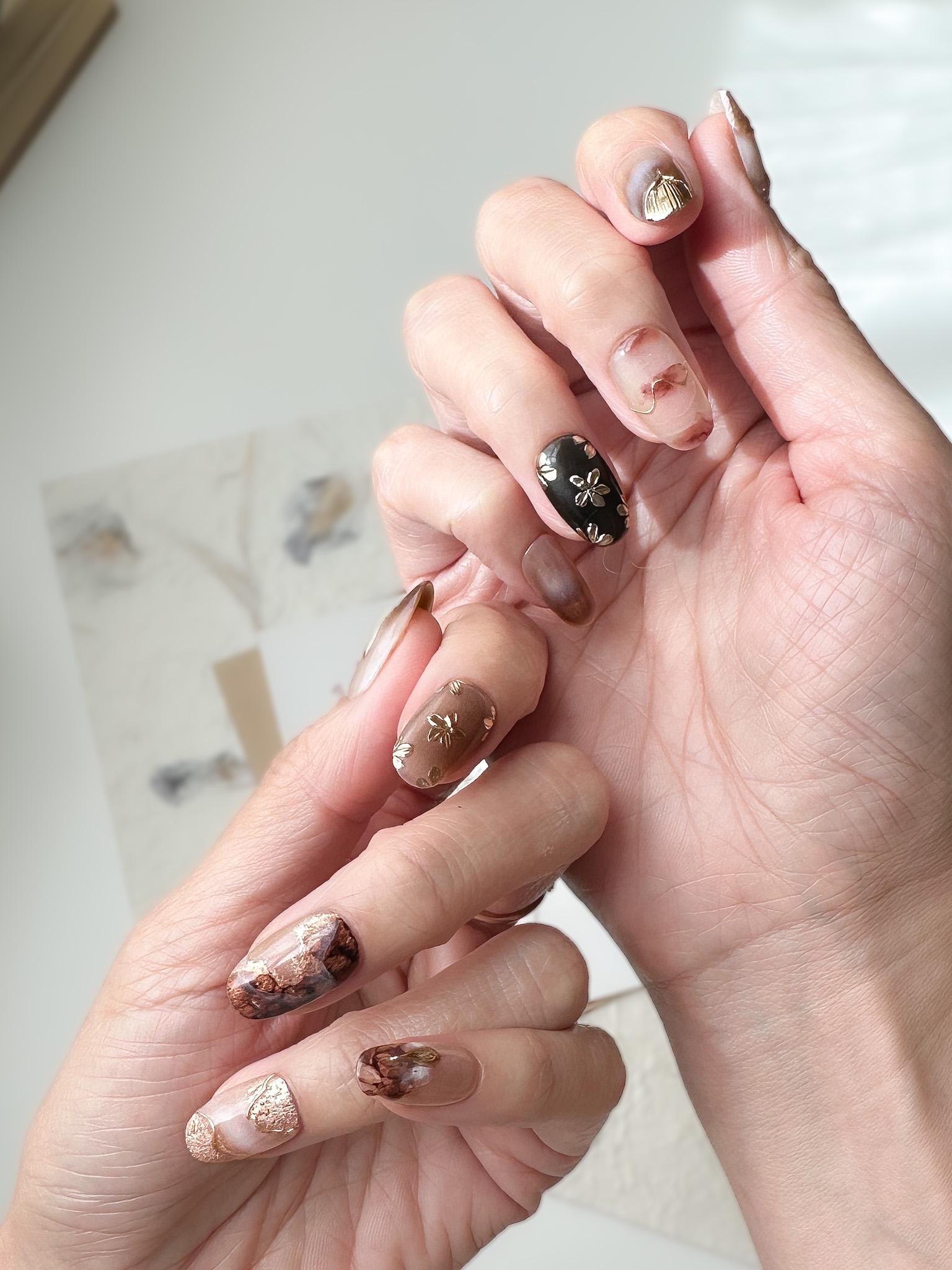 Hand with press-on nails in neutral tortoise design and mocha tones with gold foil accents, marble detailing, and embossed metallic floral designs