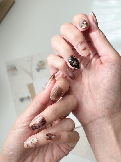 Hand with press-on nails in neutral tortoise design and mocha tones with gold foil accents, marble detailing, and embossed metallic floral designs