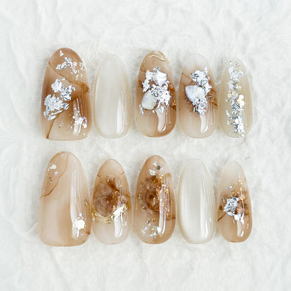 Close-up of almond-shaped nude and caramel brown press-on nails with gold foil accents and subtle shimmer