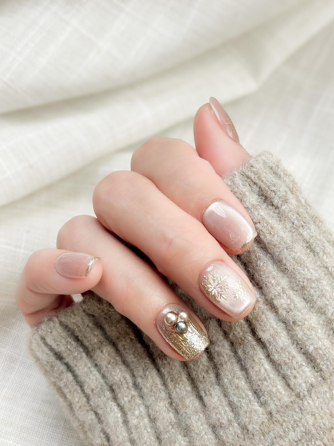 Hand with nude pink press-on nails with gold snowflake designs and glitter accents, featuring pearl embellishments for a festive holiday look