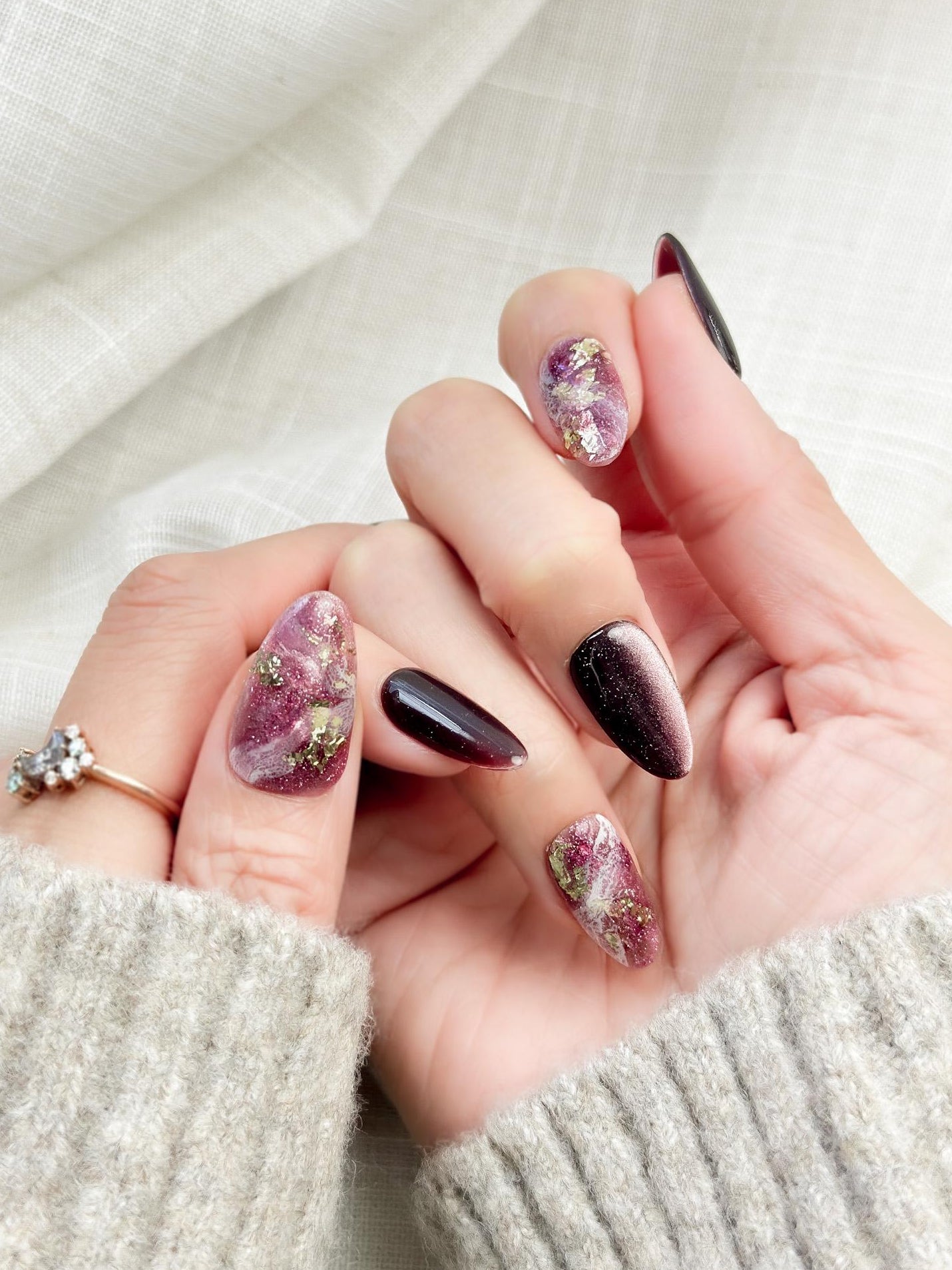 Hand with Almond-shaped press-on nails in deep plum cat-eye shimmer with pink and gold marble accents, gold foil detailing, and glossy finish