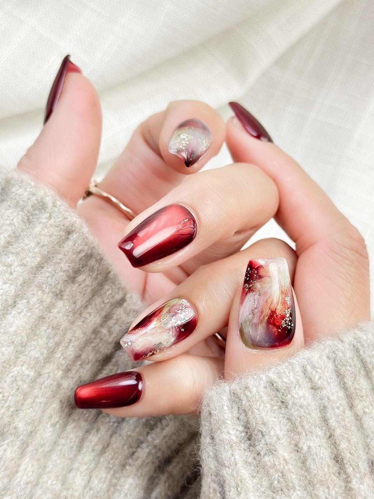 close up of hand with press-on nails in deep burgundy red with multi-color abstract art accents marble effect and silver glitter details, festive holiday style nails