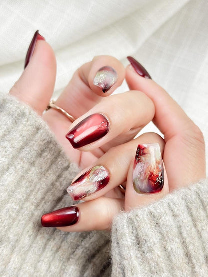 close up of hand with press-on nails in deep burgundy red with multi-color abstract art accents marble effect and silver glitter details, festive holiday style nails