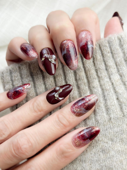 Almond-shaped burgundy and wine-red press-on nails with glossy finish, abstract marble art, gold glitter accents, and crystal pearl embellishments