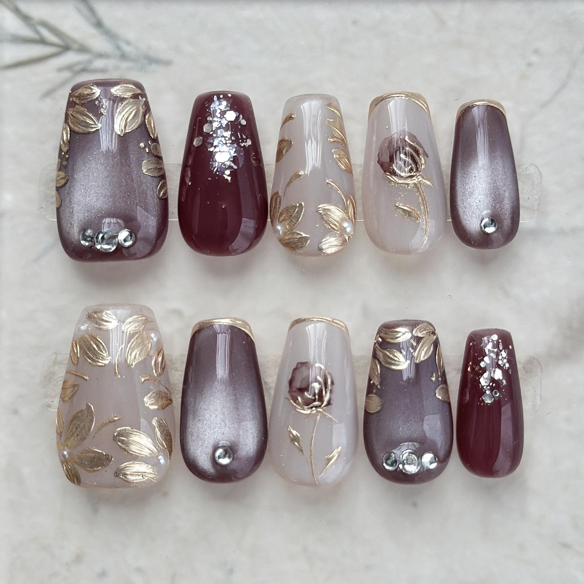Press-on nails in square coffin shape in deep plum and mauve tones with gold leaf accents, metallic floral designs, and silver gemstone details.