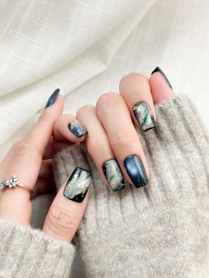 Hand with blue cat-eye press-on nails with galaxy shimmer and abstract marble accents in green, gold, and silver foil